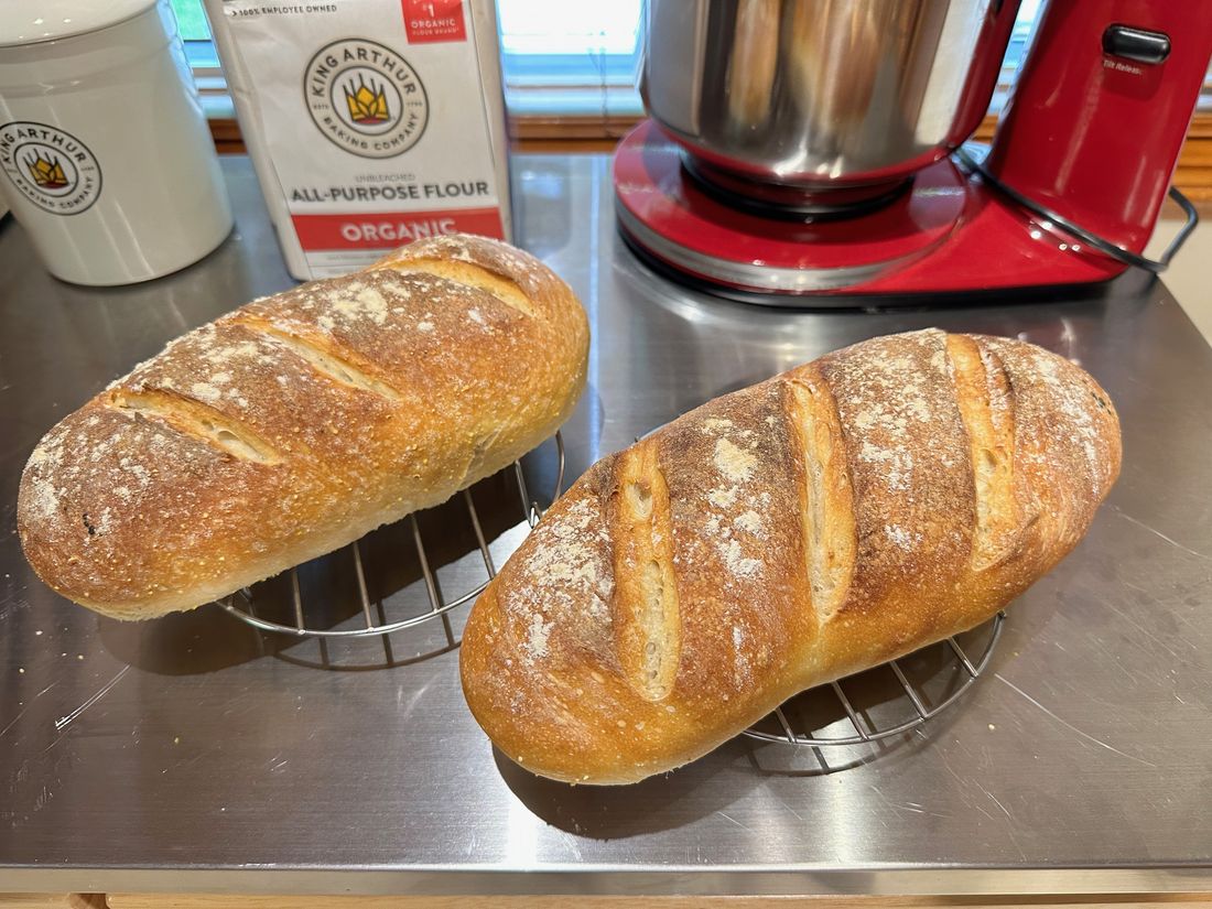 Two scored sourdough batards with golden crusts cooling on a wire rack on a steel counter.