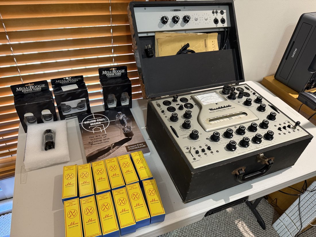 The Heathkit TT-1 mutual conductance tube tester open on a bench, its grey panel full of black knobs and tube sockets under a large meter, with boxes of new Mesa/Boogie and JJ Electronic tubes and a book on tube design beside it.