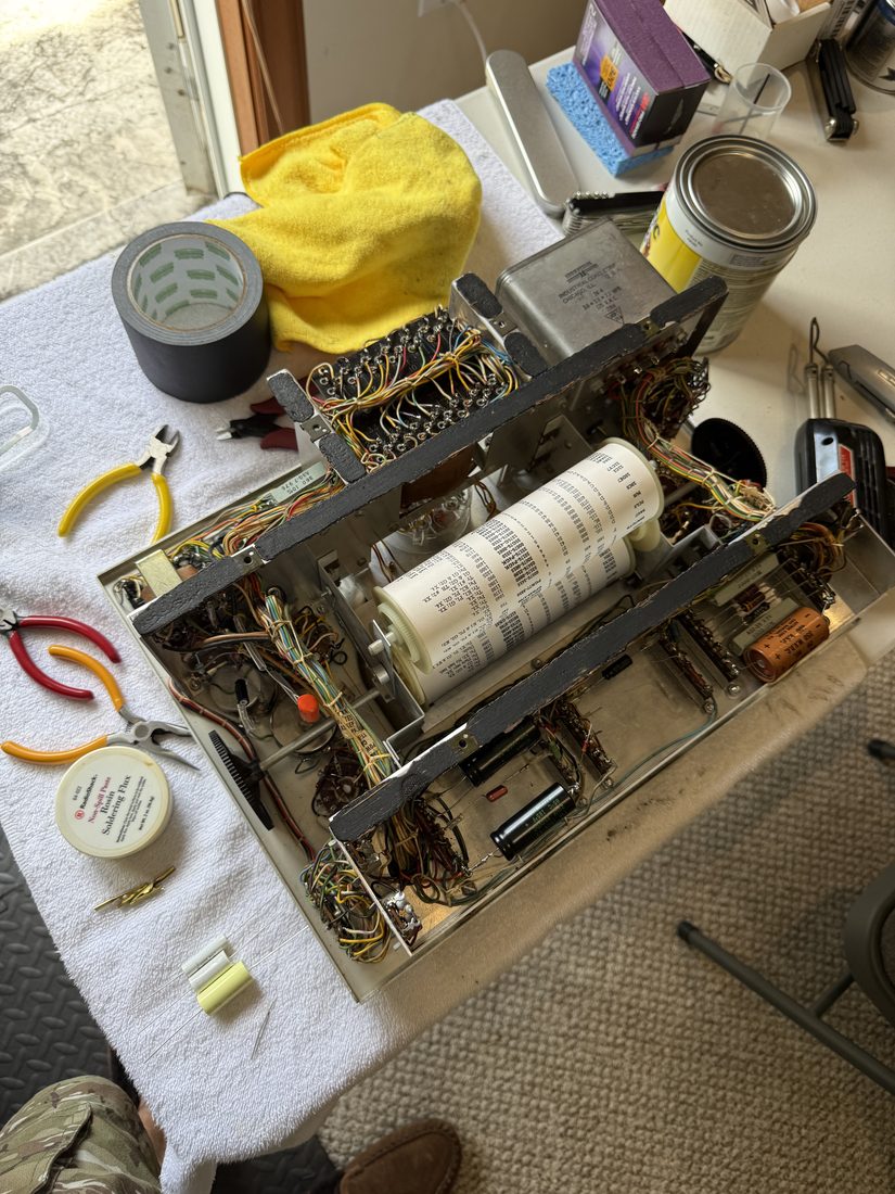 The TT-1 chassis out of its case on a towel: the multi-tap transformer's terminal board bristling with colored wires at the top, the paper roll chart of tube settings on its spool in the middle, new capacitors at the bottom, and pliers, cutters, flux, and tape around it.